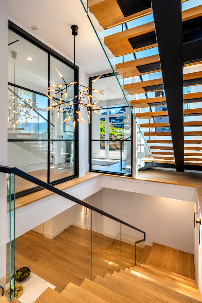 Modern floating wood staircase with glass railings, black steel supports, and a sculptural chandelier in a luxury Steamboat Springs residence.